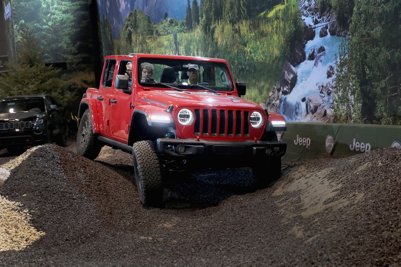 Auf dem Bild ist ein rot lackierter Jeep zu sehen, der auf einer unebenen Unterlage fährt, mit einer Naturlandschaft im Hintergrund.