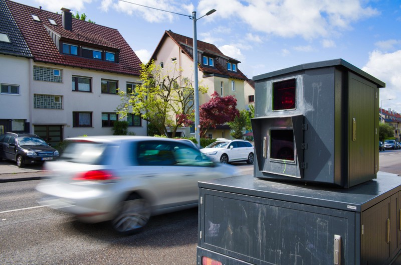 Auf dem Bild ist ein Geschwindigkeitsmessgerät zu sehen, das an einer Straße aufgestellt ist, während ein Auto vorbeifährt.