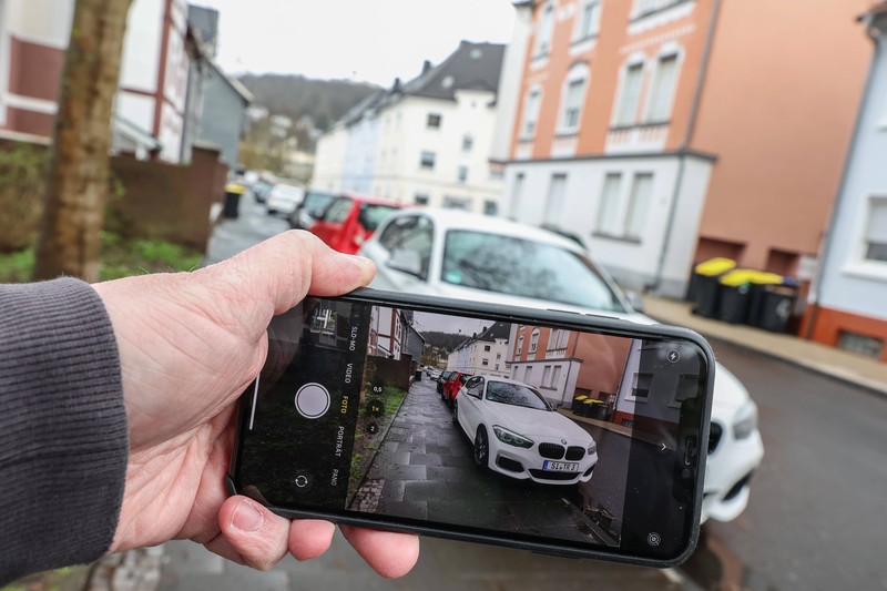Eine Person hält ein Smartphone und fotografiert ein falsch geparktes Auto auf einem Gehweg